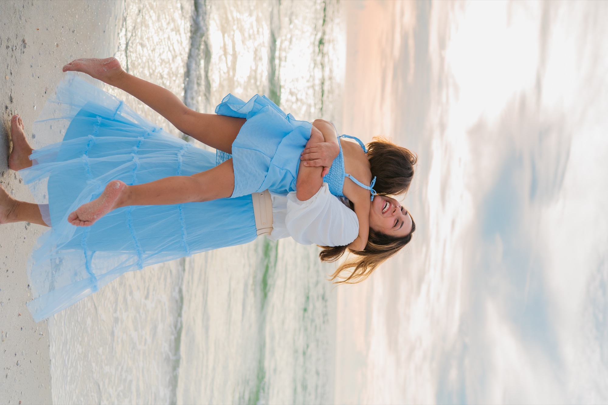A laughing mother spinning her daughter around in a hug at the water's edge on St. Pete Beach, Tampa