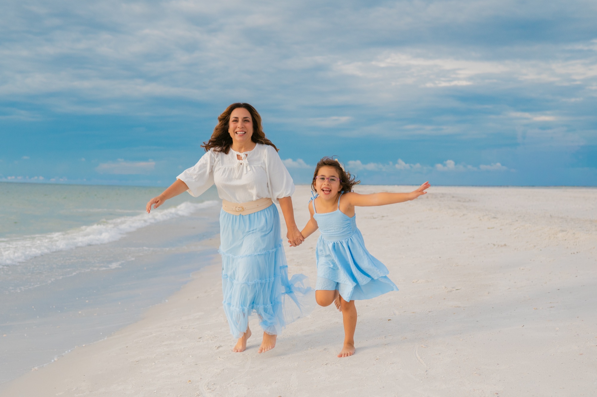 A mother and her daughter in matching blue running hand in hand along the white sand of St. Pete Beach, Tampa