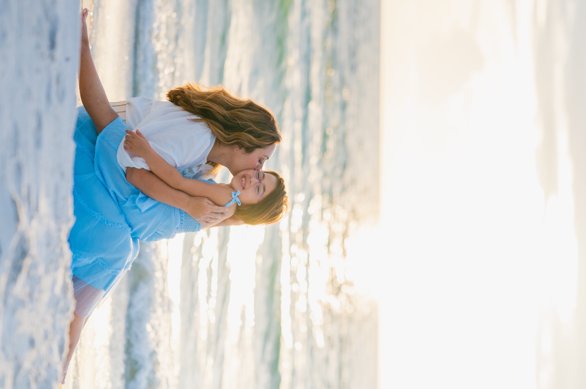 A mother kissing her daughter on the cheek as they sit on the sand at sunset, St. Pete Beach, Tampa
