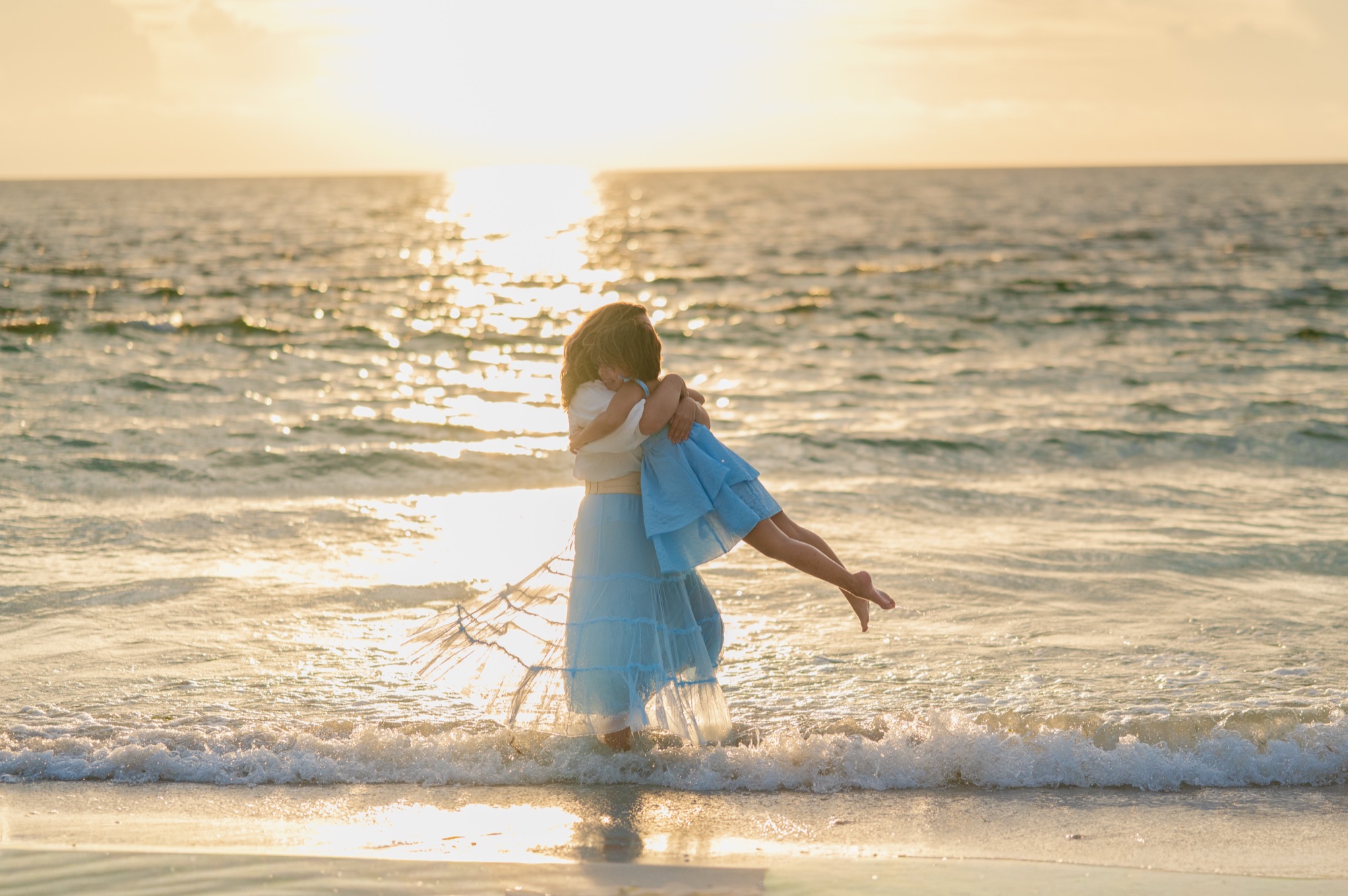 A mother lifting her young daughter in a hug in the shallow surf at sunset on St. Pete Beach, Tampa