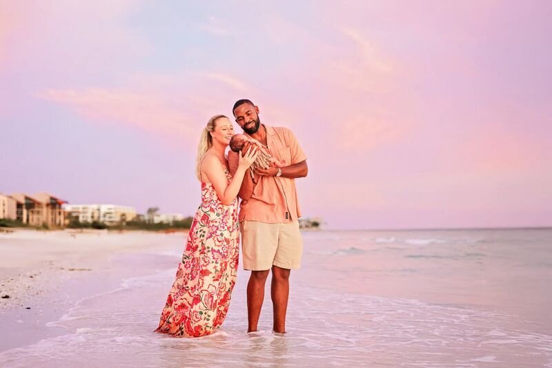 New parents cradling their newborn on the shoreline at Clearwater Beach, Tampa, during a pink and golden sunset — Tampa family photographer session by Dreamgraphy