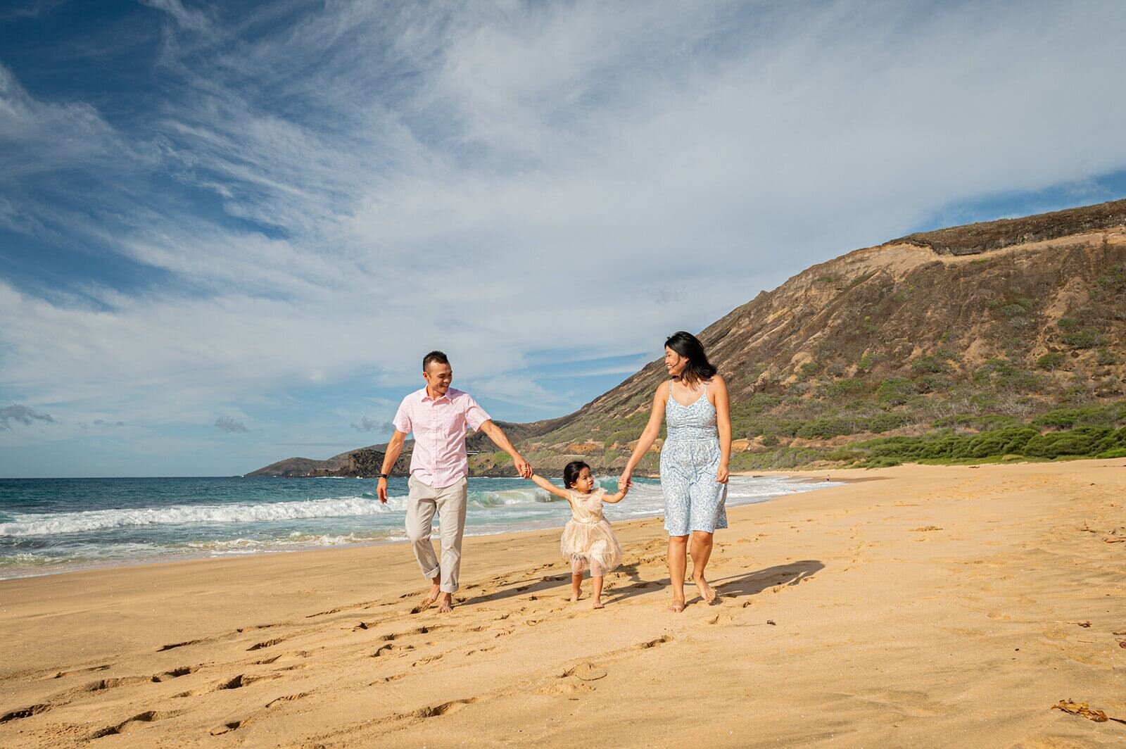 Family of three holding hands and walking along Sandy Beach Park with cliffs and crashing waves behind them