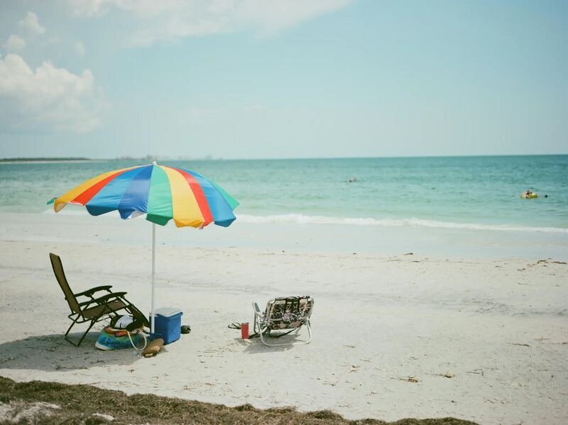A rainbow-striped beach umbrella, two beach chairs and a blue cooler on white sand beside turquoise water at Clearwater Beach