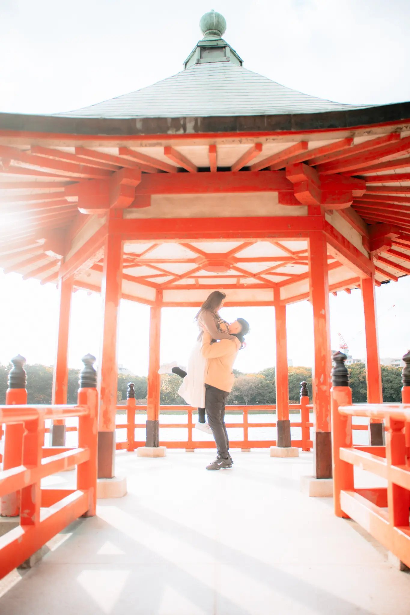 Fukuoka Couples Photography — Ohori Park Pavilion Session | Dreamgraphy A couple sharing a joyful lift inside the iconic red pavilion at Ohori Park in Fukuoka, surrounded by soft natural light and the park's serene lakeside setting. Photographed by Dreamgraphy.
