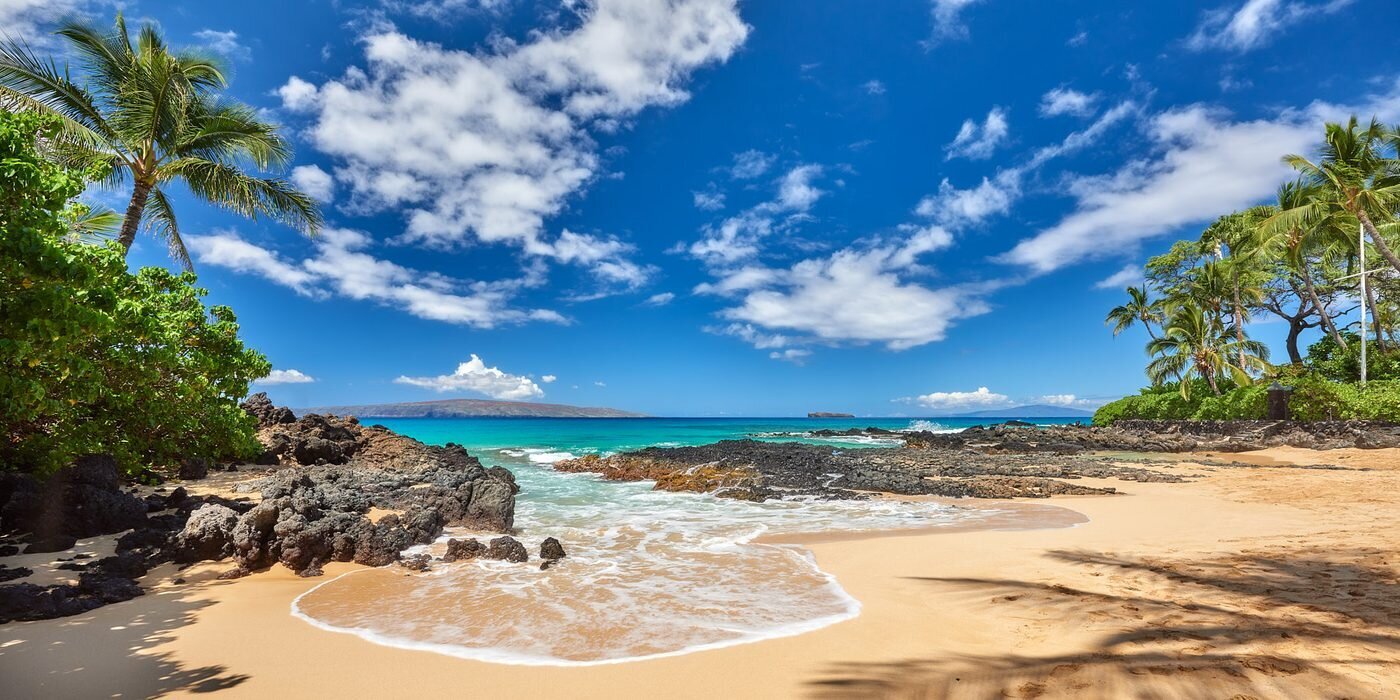 A small golden-sand cove with black lava rocks, palm trees and turquoise water under a bright blue sky on Maui