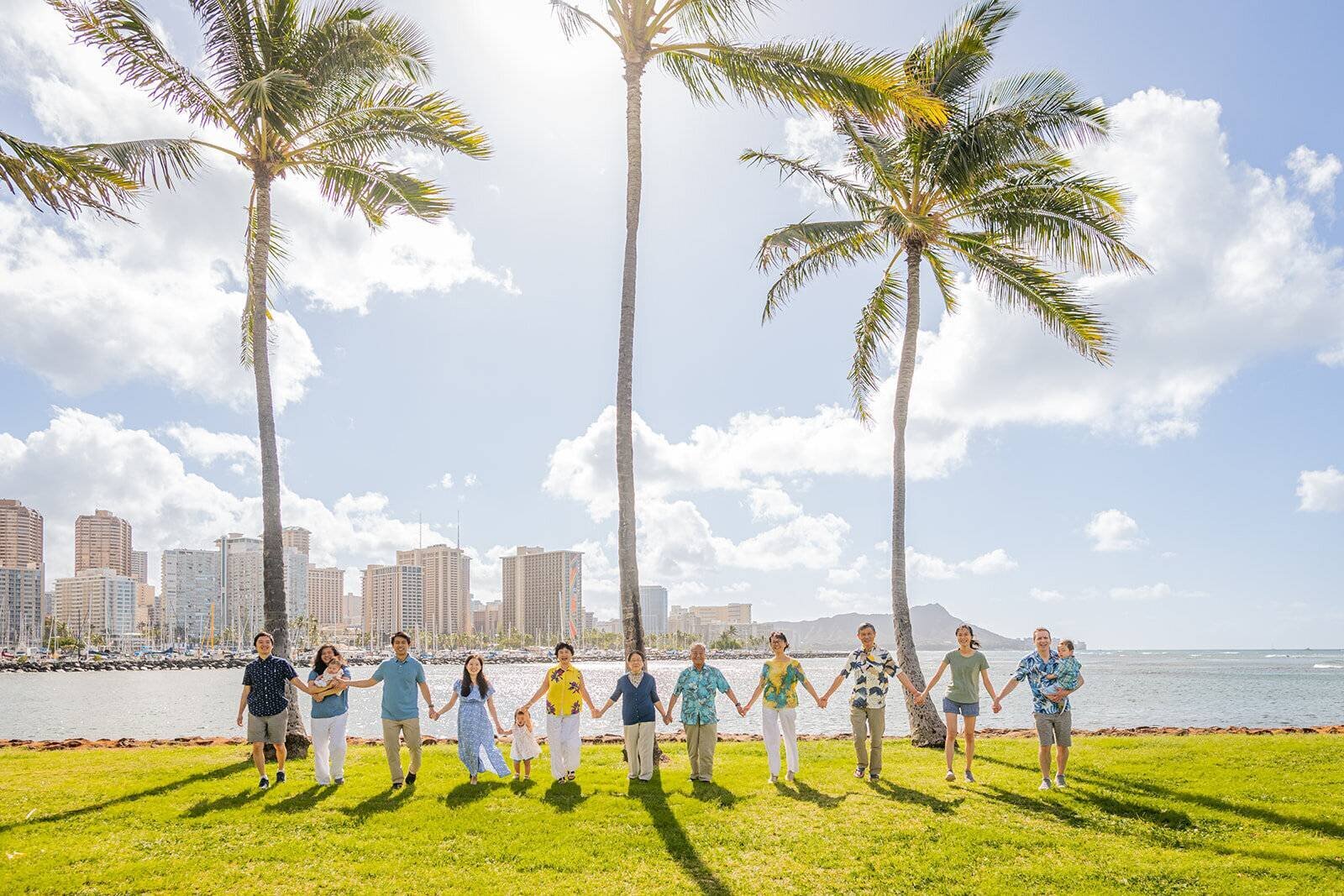 A large extended family of twelve holding hands in a line on the grass at Ala Moana Beach Park with tall palm trees, the Waikiki skyline, and Diamond Head in the background