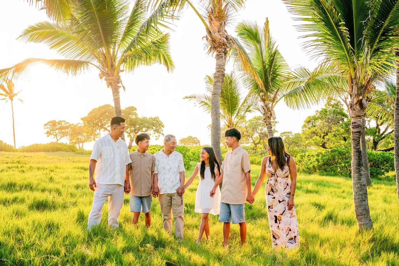 Big Island Family Photographer — Extended Family Session Under Palm Trees An extended family of six holding hands and walking together through lush green grass under tall palm trees on the Big Island of Hawaii during golden hour