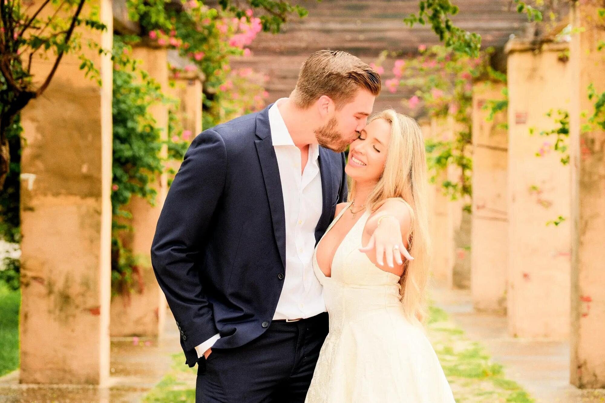 miami-engagement-photographer-vizcaya A man in a navy suit kissing the cheek of a smiling woman as she shows her ring, among stone pillars and pink bougainvillea in Miami