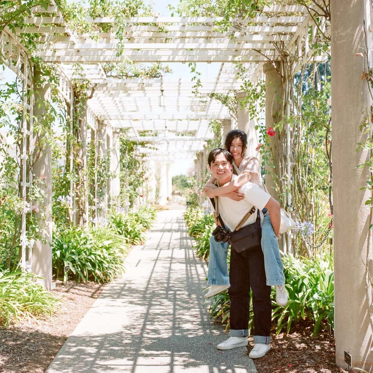 Los Angeles Couples Photographer — Couples Session at the Huntington Library Rose Garden Los Angeles couples photographer capturing a couple laughing together as the man gives his partner a piggyback ride under a white pergola covered in climbing roses at the Huntington Library Rose Garden