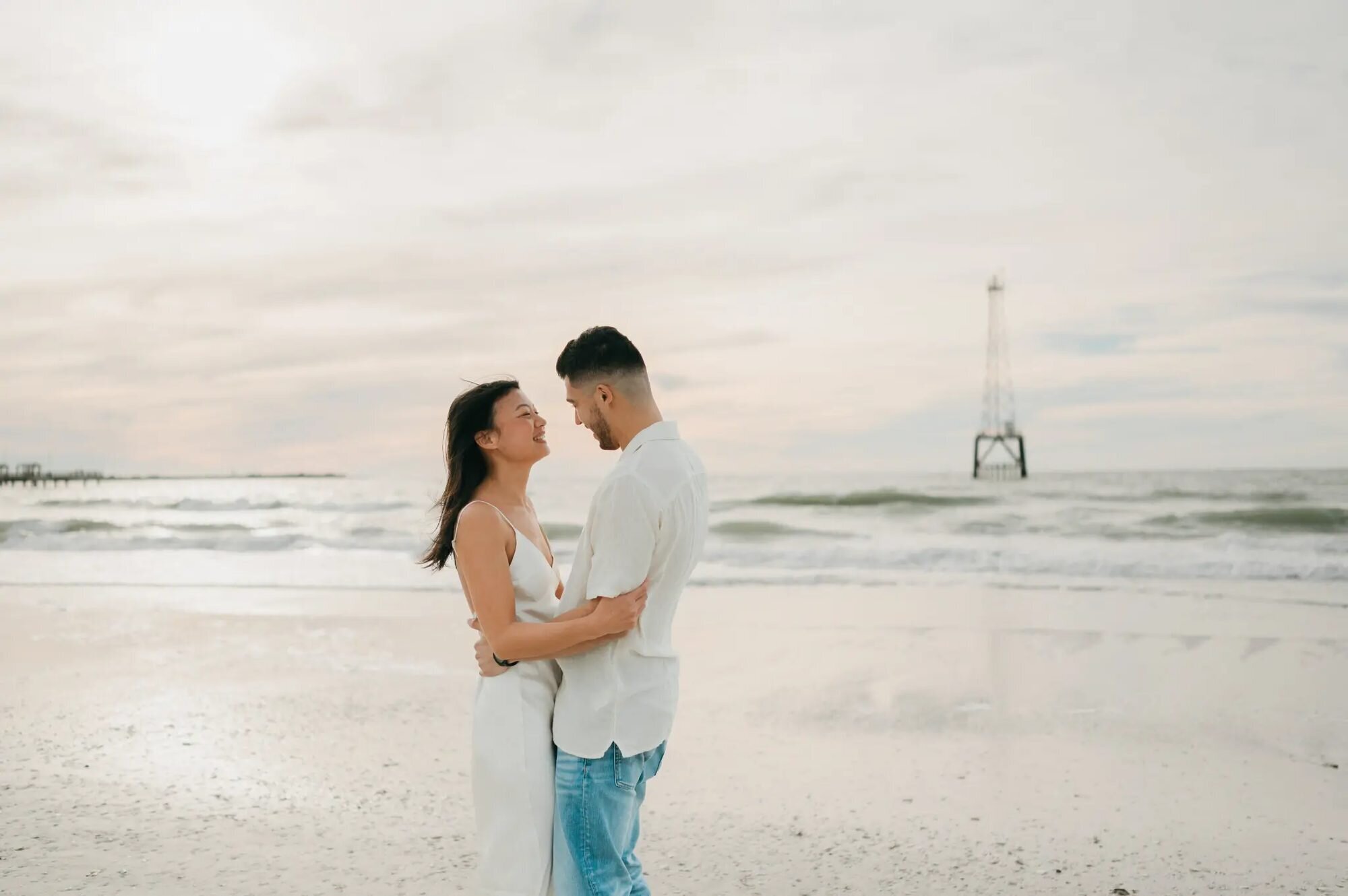 Tampa Photographer | Couple Session at Clearwater Beach A couple embracing and laughing on the white sand beach at Clearwater Beach, Tampa, with a pier and calm Gulf waters in the background — captured by a Dreamgraphy Tampa photographer