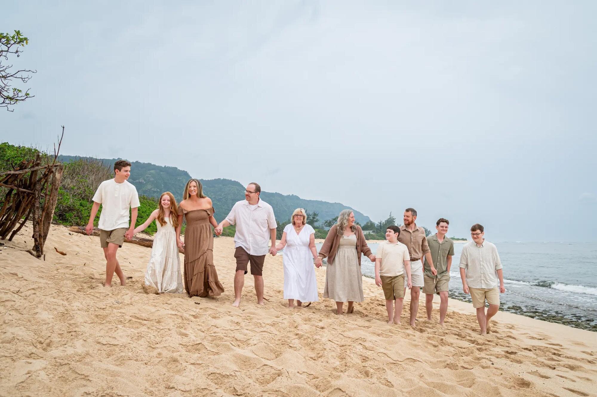 A multi-generational family of ten walking hand in hand along a sandy Kauai beach with green mountains behind