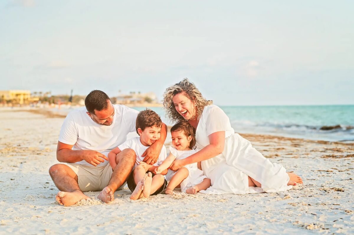 Family of four in white outfits laughing together on the white sand at St. Pete Beach, Tampa — candid family portrait by Dreamgraphy