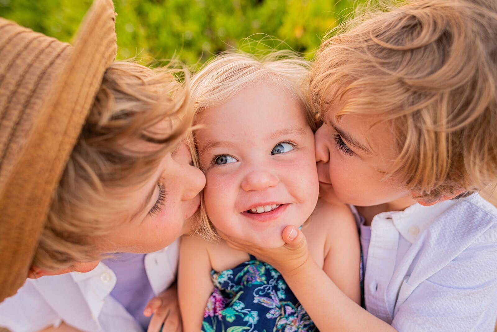 Two older siblings kissing a laughing toddler on both cheeks with lush greenery in the background