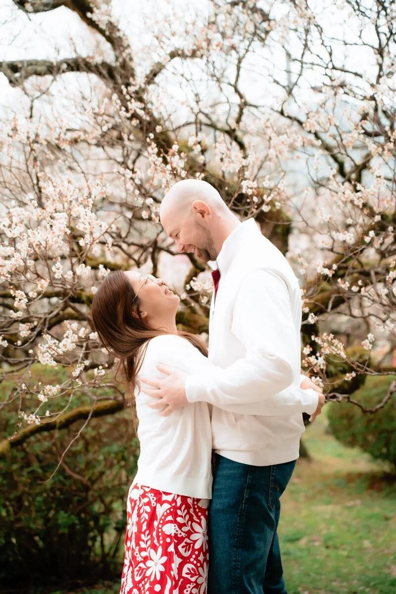 fukuoka-photographer-location-uminonakamichi-seaside-park-mobile A laughing couple in white tops embraces beneath a tree covered in pale blossoms at Uminonakamichi Seaside Park in Fukuoka.