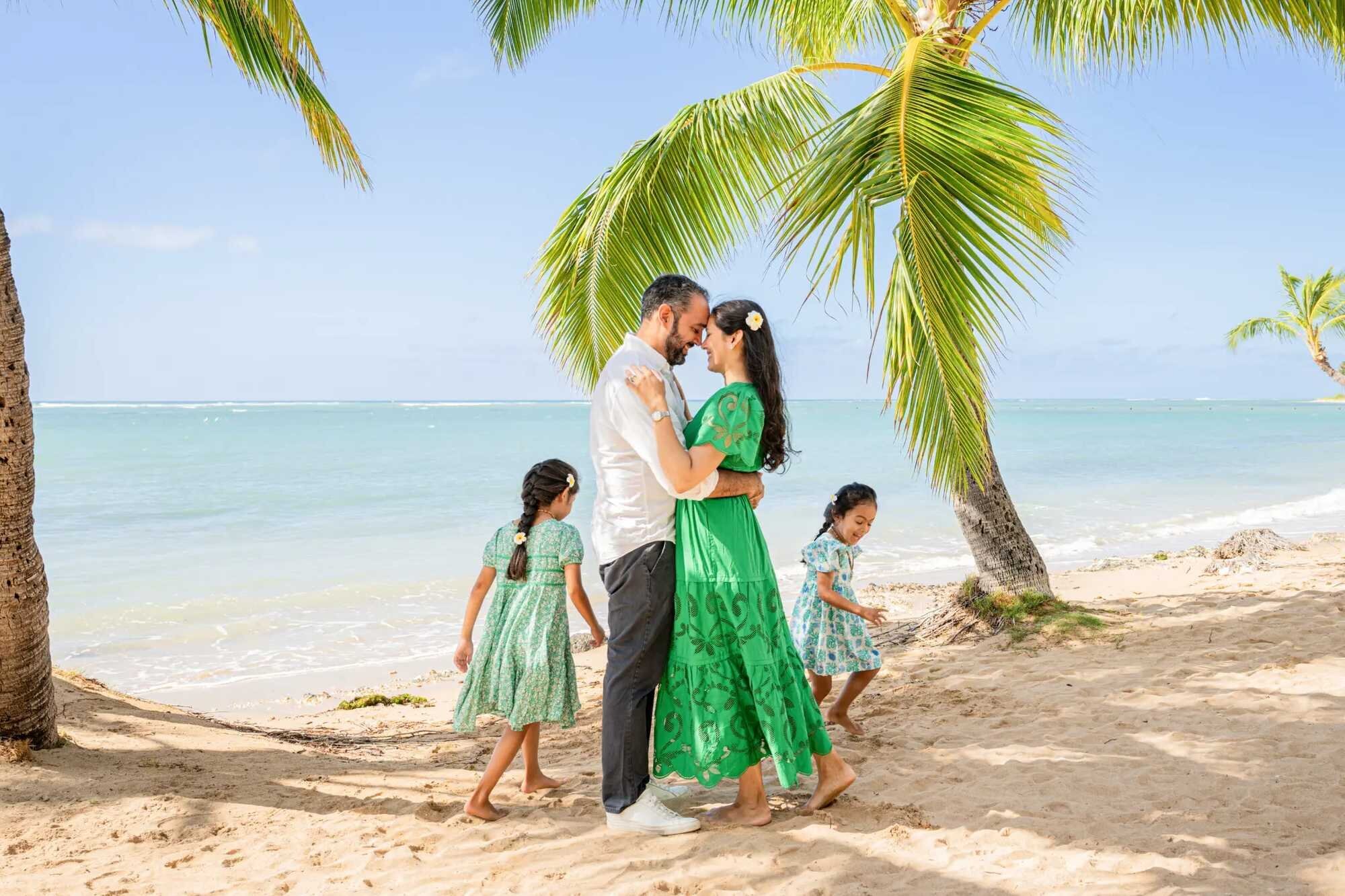 Couple embracing under a leaning palm tree at Kahala Beach while their two daughters play in the sand behind them