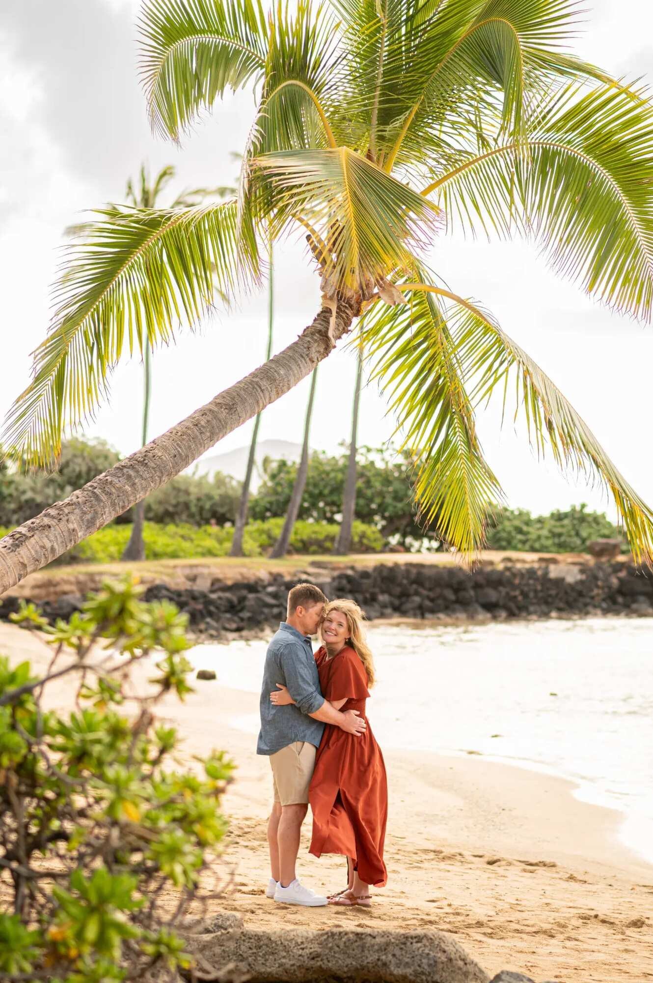 A couple embracing on a sandy Oahu beach under a tall leaning palm tree with calm ocean water and lush greenery in the background