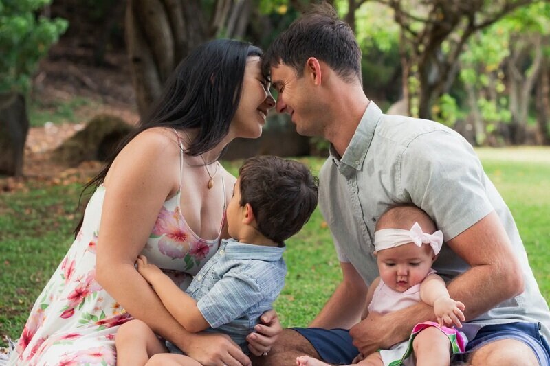 Young family of four — mom, dad, toddler son, and baby daughter — sitting together on the grass under tropical trees in Kauai, sharing a tender forehead-to-forehead moment — Kauai family photographer session by Dreamgraphy