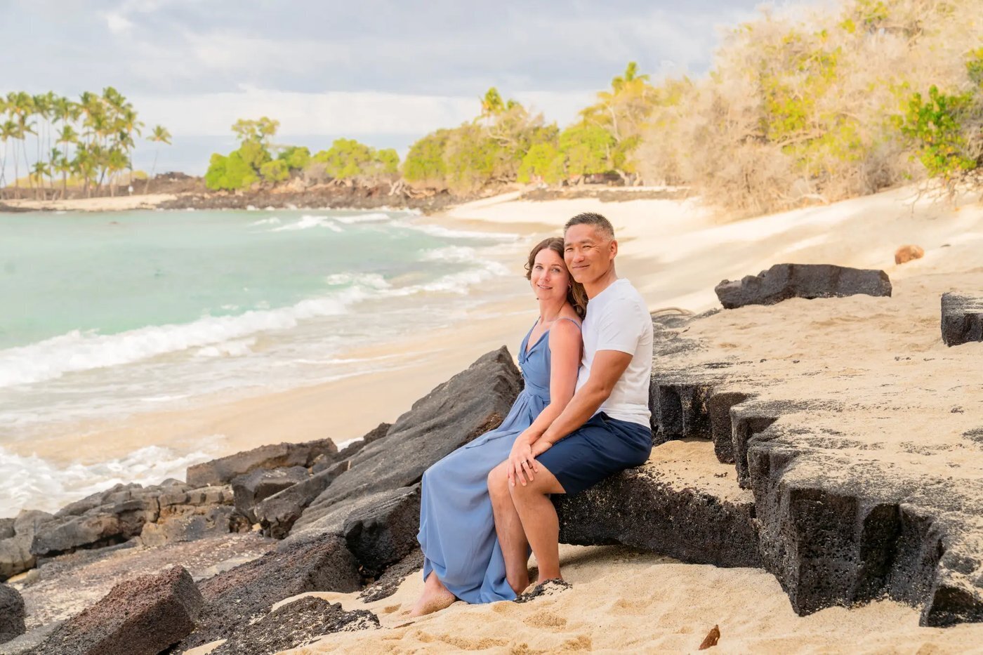 Couples Photographer Big Island Hawaii — Lava Rock Oceanside Session A couple sitting close together on lava rocks on the Big Island of Hawaii with turquoise ocean waves and a white sand beach curving behind them